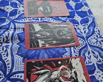 May include: A red tarot card deck and three tarot cards are displayed on a blue and white patterned cloth. The cards feature black and white illustrations with red borders. One card reads "Queen of Cups."