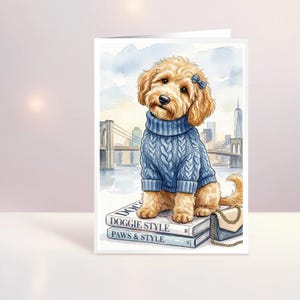 May include: A greeting card featuring a golden doodle wearing a blue cable-knit sweater and a small blue bow. The dog sits on two stacked books labeled Doggie Style and Paws & Style, next to a small handbag, with a city skyline in the background.