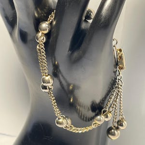 May include: A gold-toned bracelet with a delicate chain design and multiple spherical beads. The bracelet features a series of dangling chains with small beads at the ends, adding a touch of elegance. The clasp is also gold-toned.