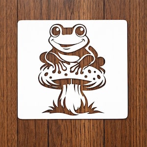 May include: A white stencil featuring a smiling frog sitting on a mushroom with dotted cap. The frog has large eyes and is surrounded by a detailed design. The stencil is on a wooden surface.