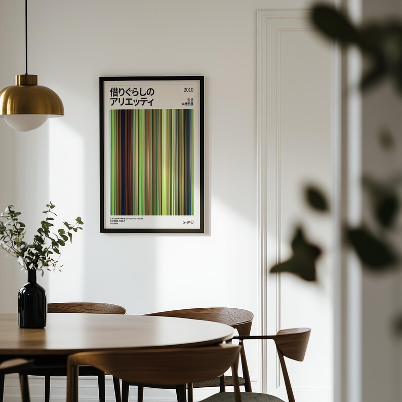 May include: A framed art print with Japanese text and vertical colour bars in shades of green, brown, and beige. The print hangs on a white wall above a wooden table and chairs. A black vase with greenery and a gold pendant light are also visible.