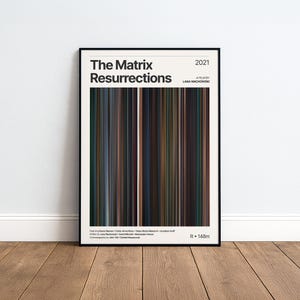 Peut inclure: Une affiche encadrée pour le film "The Matrix Resurrections", avec un design de barres de couleurs verticales. L'affiche est dans un cadre noir et affiche le titre du film, l'année 2021 et le nom du réalisateur. Elle est présentée sur une surface en bois.