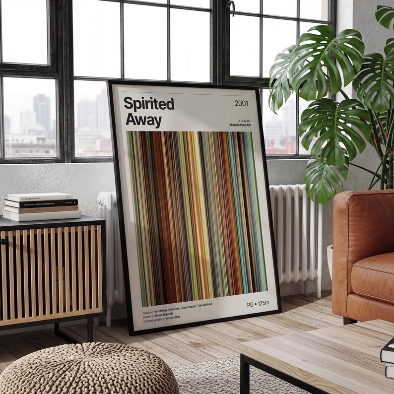 May include: A framed poster featuring the title "Spirited Away" with a colorful vertical stripe design. The poster includes the year 2001 and other text. The artwork is displayed in a living room setting with a window, a wooden cabinet, and a leather sofa.
