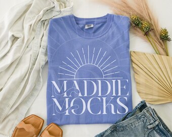 May include: A blue t-shirt with a white sun and the text "MADdie MOCKS" printed on the front.