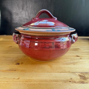 May include: A round ceramic lidded pot with a glossy deep red glaze. The pot features two side handles and a matching lid with a central handle. A beige band encircles the lid, decorated with small, dark teal leaf-like patterns.