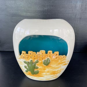 May include: A white ceramic vase with a unique cutout design. The vase features a desert landscape scene with a yellow and orange base, a teal interior, and a cityscape design. Green cactus and heart-shaped details add to the aesthetic.