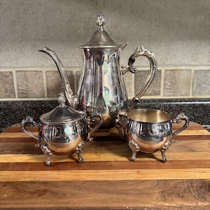 May include: A silver-plated tea service set consisting of a tall teapot with a lid, a lidded sugar bowl, and an open creamer. Each piece features a polished metallic finish, ornate curved handles, and decorative feet with intricate detailing.