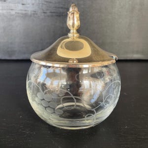 May include: A clear glass jar with a silver lid and a gold-colored finial. The jar features an etched grape design. The jar is round and sits on a dark surface. The lid has a curved shape.