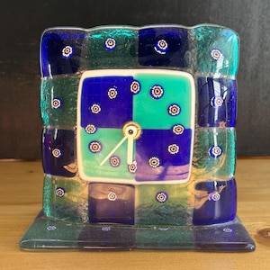 May include: A square glass clock featuring a checkered pattern of deep blue and teal squares. The clock face is adorned with small, circular floral-like millefiori glass accents in white, red, and blue. It has two simple white clock hands.