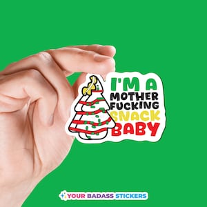 May include: A white sticker with a Christmas tree design and the text "I'M A MOTHER FUCKING SNACK BABY" in green, yellow, and red. The sticker is held against a green background.