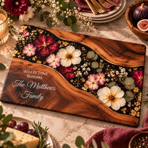 Op de afbeelding: Een rechthoekige houten serveerplank met een golvende harsinleg in het midden, gevuld met geperste witte, roze en donkerrode bloemen en groen. Het hout is gegraveerd met de witte tekst "WHERE LOVE BLOOMS" en "The Matthews Family".