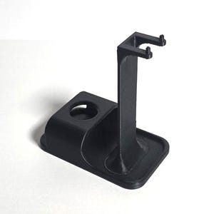 May include: A black, 3D-printed shaving stand. The stand features a tall, vertical post with a curved top designed to hold a razor. It also has a circular holder and a small base. The stand is made of a matte black material.