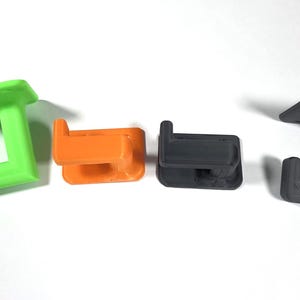 May include: Four plastic wall hooks in green, orange, dark gray, and gray. Each hook has a flat top with screw holes and a J-shaped hook extending downwards. The hooks are designed for hanging items.