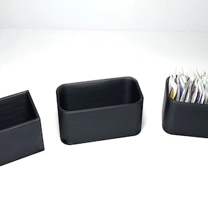 May include: Three black rectangular containers of varying sizes. One is angled, one is a simple box, and the third is filled with packets. The containers are made of a matte black material and are set against a white background.