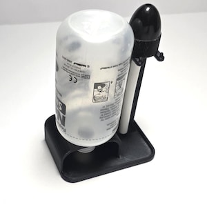May include: A black plastic stand holding a white bottle and a black and white device. The bottle has a white label with black text. The device has a black top and a white body. The stand is on a white surface.