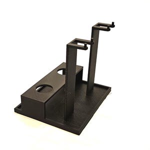 Puede incluir: Un soporte de plástico negro con dos soportes verticales y una base con dos recortes circulares. El soporte está diseñado para sostener objetos en posición vertical.