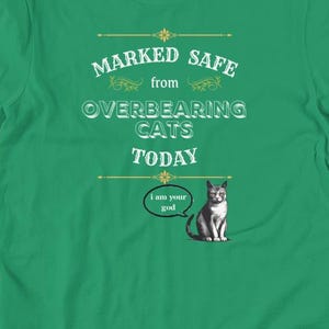 Puede incluir: Una camiseta verde con el texto "MARKED SAFE from OVERBEARING CATS TODAY". Incluye una ilustración de un gato con el bocadillo "I am your god".
