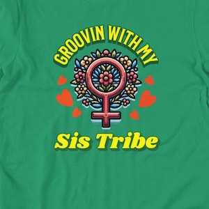 Può includere: Una maglietta verde con la scritta "Groovin' With My Sis Tribe" in giallo e bianco. Il design include un emblema floreale con un simbolo femminile e accenti a forma di cuore rosso.