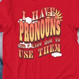 Może przedstawiać: Czerwona koszulka z napisem "I HAVE PRONOUNS AND I KNOW HOW TO USE THEM" w stylu retro. Projekt zawiera słońce, chmury i schemat kolorów tęczy. Koszulka jest prezentowana na płaskiej powierzchni z dżinsami i akcesoriami.