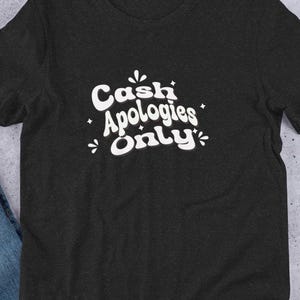 May include: Dark grey t-shirt featuring the white text "Cash Apologies Only" in a whimsical font. The short-sleeved shirt is made of a soft material. The shirt is displayed on a flat surface with other accessories.