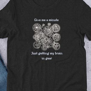 May include: Dark gray t-shirt featuring a detailed illustration of interlocking gears in shades of gray and white. The text "Give me a minute, Just getting my brain in gear" is printed above and below the gears.