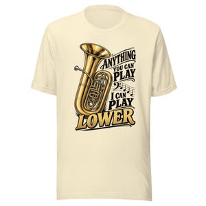 Może przedstawiać: Kremowy t-shirt z krótkim rękawem z grafiką przedstawiającą złotą tubę oraz czarny napis "ANYTHING YOU CAN PLAY I CAN PLAY LOWER" wraz z kluczem basowym i nutami, umieszczony centralnie na przodzie odzieży.