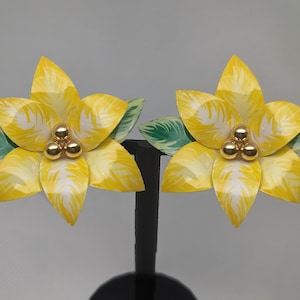 May include: A pair of flower-shaped earrings. Each earring features yellow petals with white accents, gold-colored center beads, and green and white leaves. The earrings are displayed on a black stand.