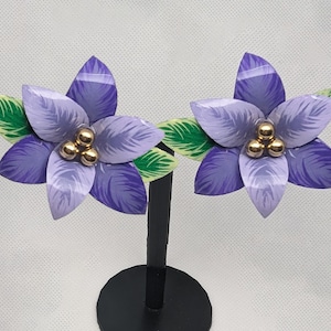May include: A pair of floral earrings featuring purple petals with gold-colored accents. The earrings have a flower design with three gold beads in the center and green leaves. The earrings are displayed on a black stand.