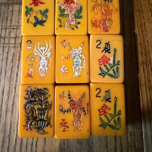 Puede incluir: Un conjunto de nueve fichas de mahjong rectangulares de color amarillo dispuestas en una cuadrícula de tres por tres. Cada ficha presenta grabados de flores, jarrones y caracteres chinos pintados en rojo, verde, negro y blanco, con números.
