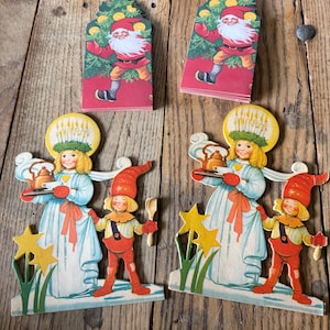 May include: Two stacks of rectangular cards featuring a gnome in a red hat and green tree, and two die-cut paper figures depicting a person in a white gown with a candle crown and a small gnome in a red hat holding a wooden spoon and tray.