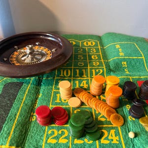 Puede incluir: Una ruleta marrón con un centro plateado se encuentra sobre un tapete de apuestas de fieltro verde con líneas de cuadrícula y números amarillos. Varias pilas de fichas de juego redondas de plástico rojo, verde, amarillo, naranja y negro.