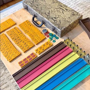 Puede incluir: Un juego de Mahjong que incluye numerosas fichas amarillas con caracteres grabados en negro y rojo, cuatro soportes rectangulares largos en marrón, rosa, amarillo, azul y turquesa, una colección de fichas de conteo de plástico y un estuche con estampado de piel de serpiente.