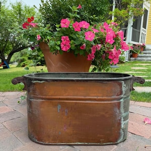 May include: An oval copper planter with a weathered patina and green oxidation marks. The vessel features two decorative metal handles on the sides and a horizontal ribbed band running along the upper portion of the exterior body.