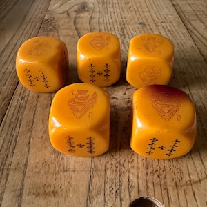 May include: Five amber-coloured poker dice featuring engraved playing card symbols. Each die displays a king, queen, jack, ten, nine, or ace, with black and red ink detailing the suits and face card imagery on the rounded, smooth surfaces.