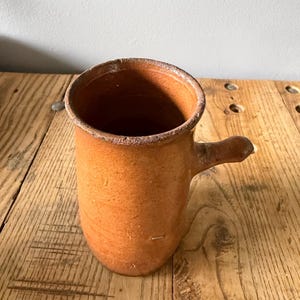 May include: A cylindrical terracotta mug with a rough, unglazed surface in a warm orange-brown tone. The vessel features a single small, protruding handle on the side and a thick, slightly uneven rim, showcasing a natural, earthy texture throughout.