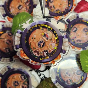 May include: A clear acrylic keychain with a purple rim and a gold glitter background. The keychain features a cartoon illustration of a pot of soup with five cartoon characters inside. The characters are all wearing different coloured hats and have different facial expressions.
