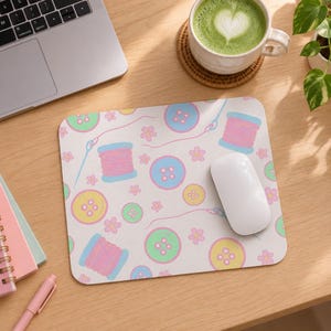 Puede incluir: Una alfombrilla de ratón rectangular con un patrón repetitivo de botones de costura en tonos pastel, carretes de hilo rosa y pequeñas flores sobre un fondo claro. Un ratón de ordenador blanco descansa sobre la alfombrilla.