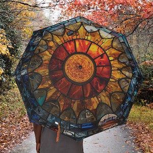 Puede incluir: Un paraguas octogonal con un diseño de vidriera que representa un sol radiante en tonos naranja, amarillo y rojo. Los bordes están delineados en azul oscuro y marrón. El fondo muestra a una persona caminando por un camino.