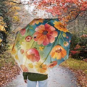 May include: An open umbrella with a vibrant floral design featuring orange, yellow, and red flowers against a teal and green background. The umbrella is held by a person walking on a path surrounded by autumn foliage.