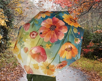 Può includere: Un ombrello aperto con un vivace design floreale con fiori arancioni, gialli e rossi su uno sfondo verde acqua e verde. L'ombrello è tenuto da una persona che cammina su un sentiero circondato da un fogliame autunnale.
