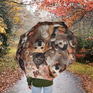 Puede incluir: Un paraguas abierto con un diseño de perros y gatos sonrientes, mariposas y flores. El paraguas lo sostiene una persona en un camino rodeado de árboles con follaje otoñal.