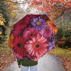 Op de afbeelding: Een open paraplu met een levendig bloemmotief van roze, rode, paarse en oranje gerbera's. Het patroon van de paraplu contrasteert met een wandelpad en herfstbladeren, wat een opvallend contrast creëert.