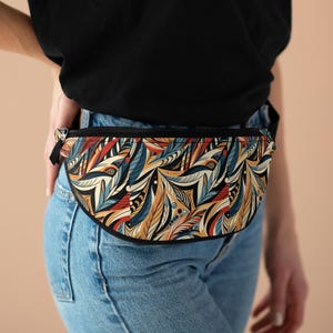 May include: A black fanny pack with a colorful, abstract feather pattern in shades of blue, red, yellow, and white. The fanny pack is worn around the waist of a person wearing blue jeans.