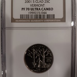 Peut inclure: Une pièce de monnaie 2001 S Clad 25C Vermont PF 70 Ultra Cameo, dans un boîtier de protection. La pièce présente un motif détaillé avec des arbres et une figure. Le boîtier contient une étiquette avec des informations de classement et un code-barres.
