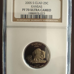 Peut inclure: Une pièce de monnaie Kansas de 2005, dans un étui de protection. La pièce représente un bison détaillé et le texte "KANSAS 1861". L'étui comprend le texte "2005 S CLAD 25C" et "PF 70 ULTRA CAMEO".