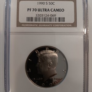 Peut inclure: Une pièce de monnaie Kennedy de 50 cents de 1990 S, classée PF 70 Ultra Cameo, dans un boîtier de protection transparent. La pièce présente un profil de John F. Kennedy et l'inscription "IN GOD WE TRUST". Le boîtier comprend un code-barres et le logo Numismatic Guaranty Corporation.