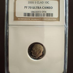 Peut inclure: Une pièce de monnaie 2005 S Clad 10C, classée PF 70 Ultra Cameo, dans un boîtier en plastique transparent. La pièce représente un profil d'homme et l'année 2005. Le boîtier porte l'inscription "NUMISMATIC GUARANTY CORPORATION".