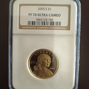 Peut inclure: Une pièce de monnaie dorée de 2005 S $1, classée PF 70 Ultra Cameo, dans un boîtier de protection. La pièce présente un portrait et l'inscription "LIBERTY". Le boîtier comprend une étiquette avec le logo Numismatic Guaranty Corporation et le numéro de série.