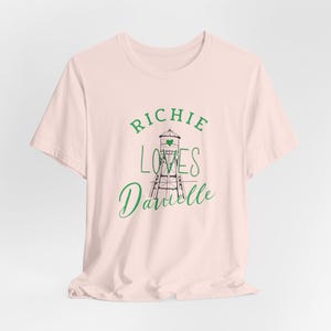 May include: A light pink t-shirt with the text "RICHIE LOVES Danielle" in green script. The design includes a green heart and a black and green illustration of a water tower. The shirt is made of a soft material.