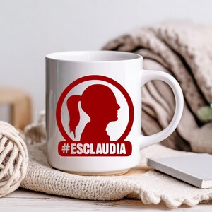 May include: White ceramic mug with a red circle design featuring a silhouette of a person with a ponytail. The text "#ESCLAUDIA" is printed in red below the circle.